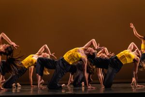 Dancers in yellow and black costumes perform a synchronized backbend on stage.