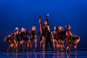 A group of dancers in dark costumes perform a synchronized routine against a blue backdrop.