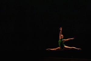 A solo dancer executes a powerful split jump silhouetted against a black background.