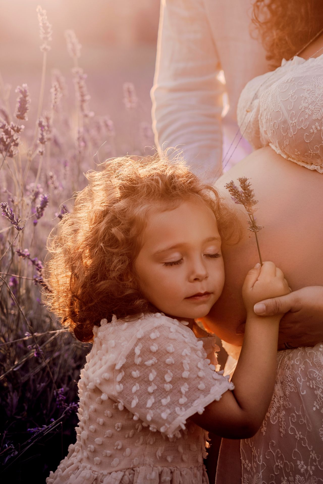 Curly-haired little girl rests her cheek on her mother's pregnant belly in a lavender field at golden hour during a maternity photoshoot.