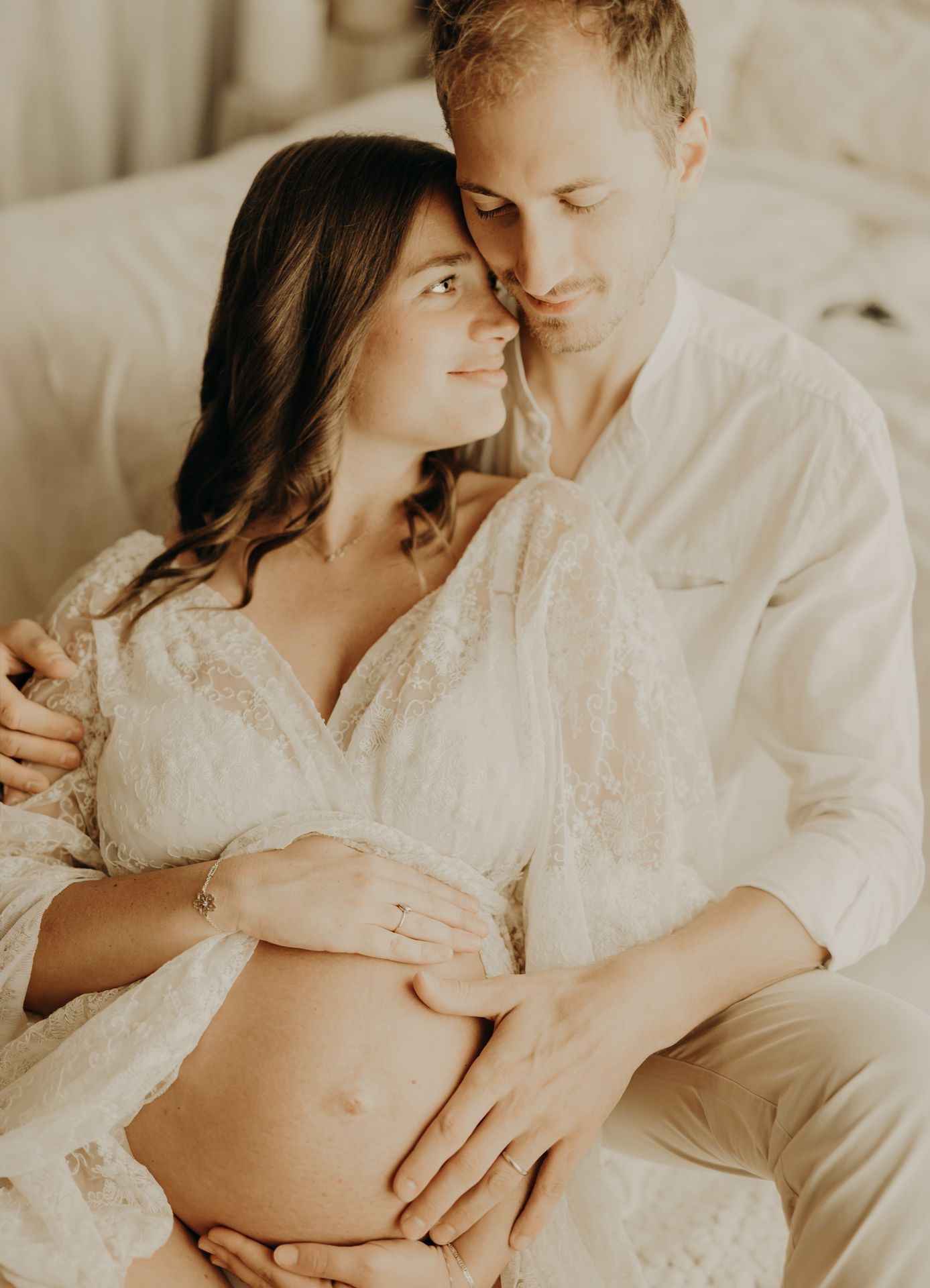 Expectant couple embracing tenderly as the man rests his hands on the woman's baby bump in a soft, light-filled maternity photo.