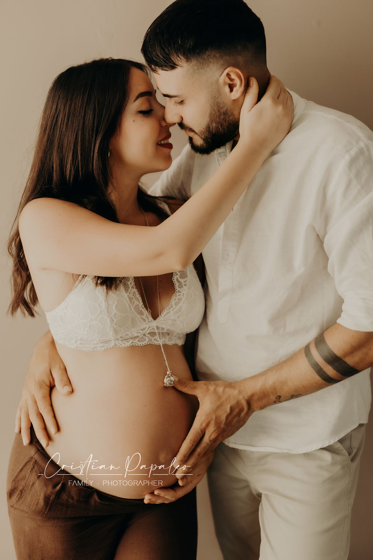 Pregnant woman in lace bralette and her bearded partner share a tender moment, touching foreheads in a warm studio maternity portrait.
