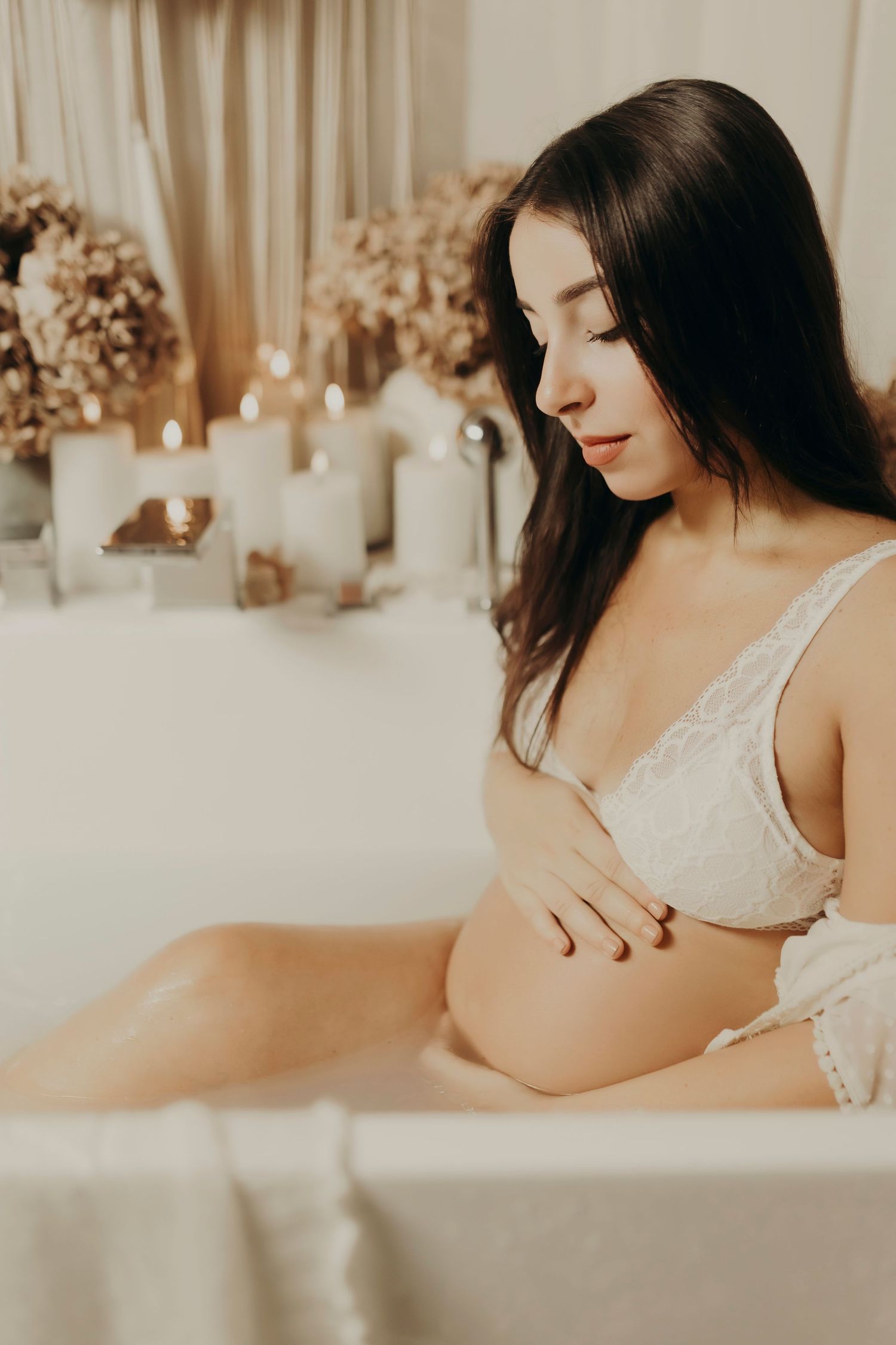 Pregnant woman in a white lace bralette cradles her bare belly while sitting in a milk bath surrounded by candles and dried flowers.