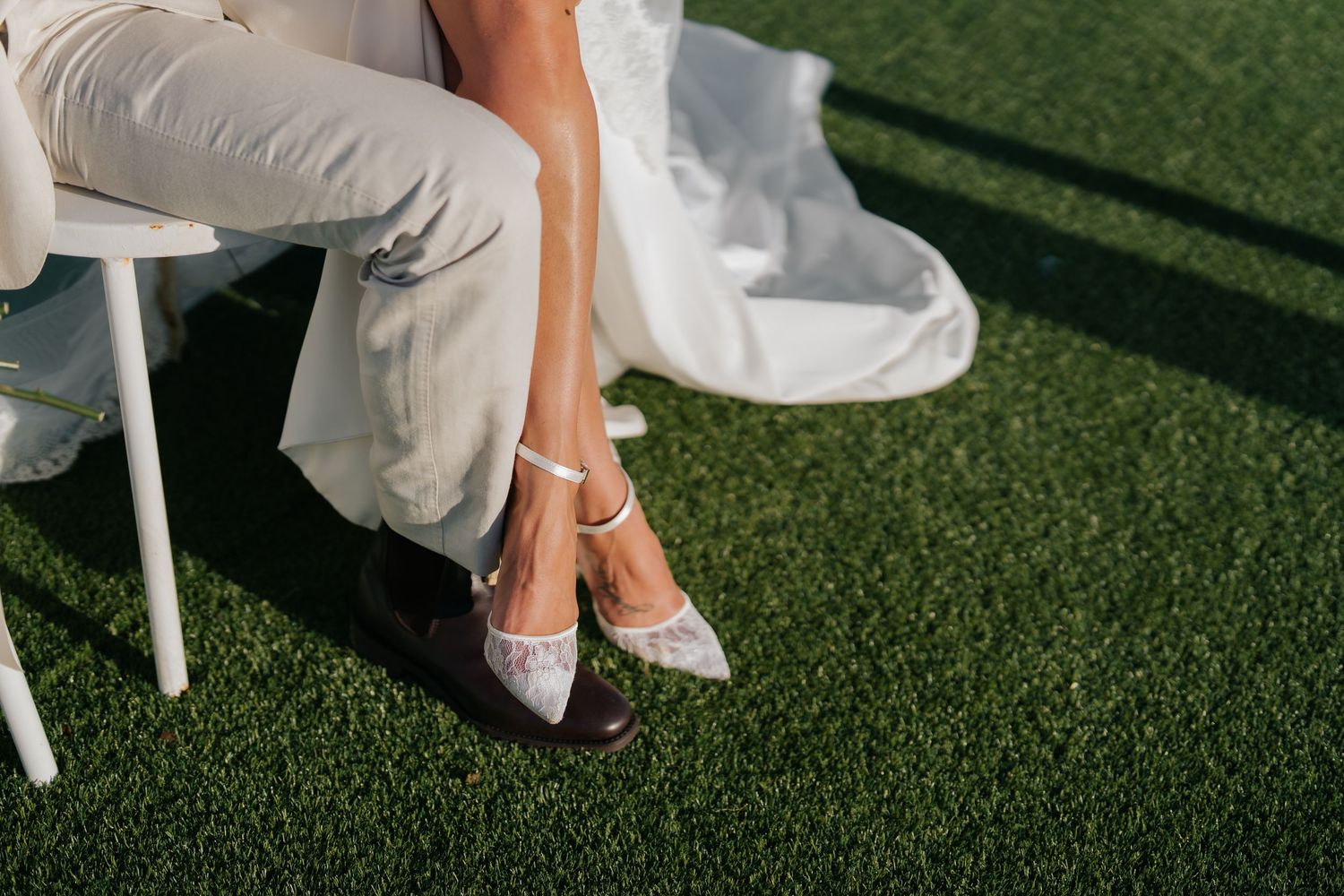 Close-up of bride and groom sitting side by side showing wedding shoes and suit trousers on grass.