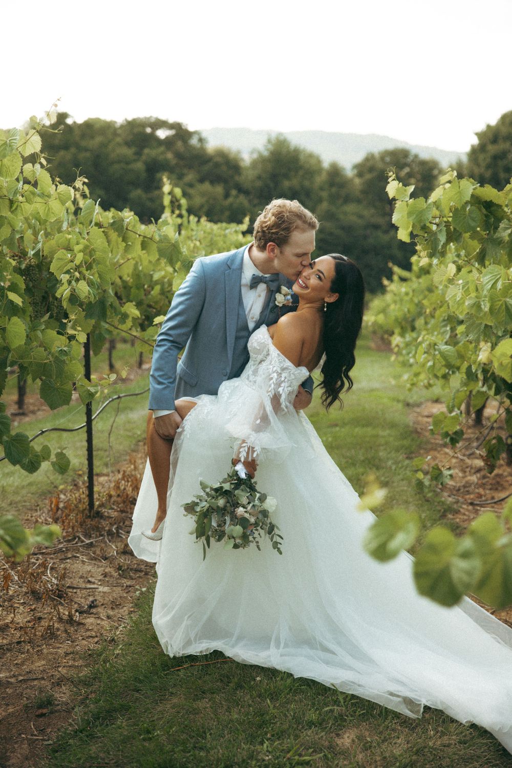 Romantic couple pose together in a vineyard with mountains in the background.