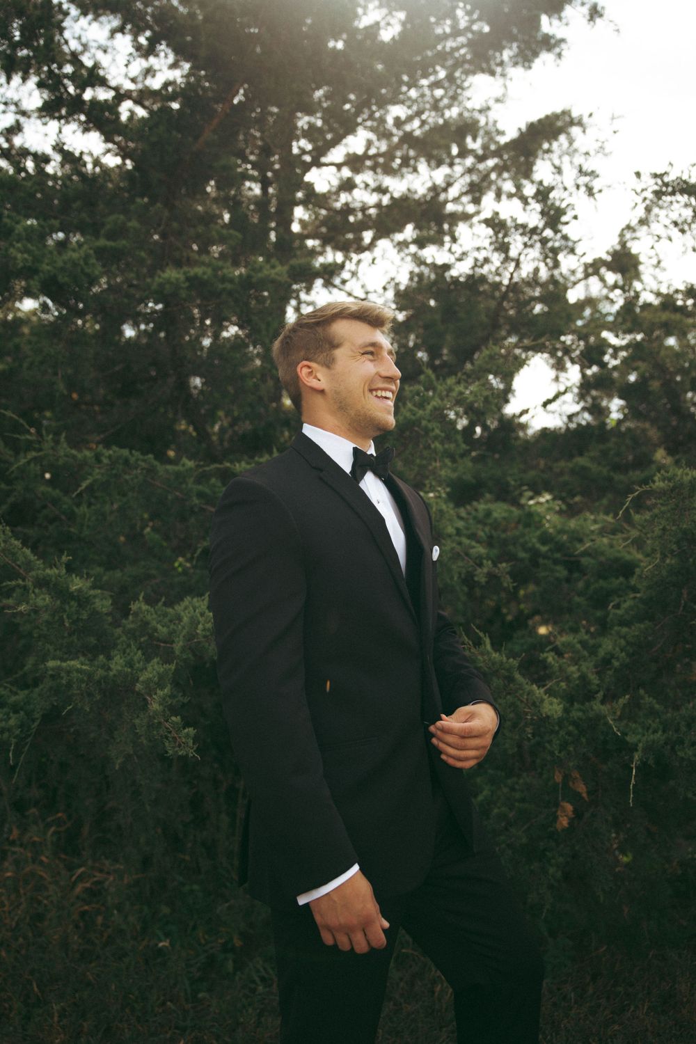 A groom in a classic black tuxedo smiles while standing outdoors surrounded by trees.