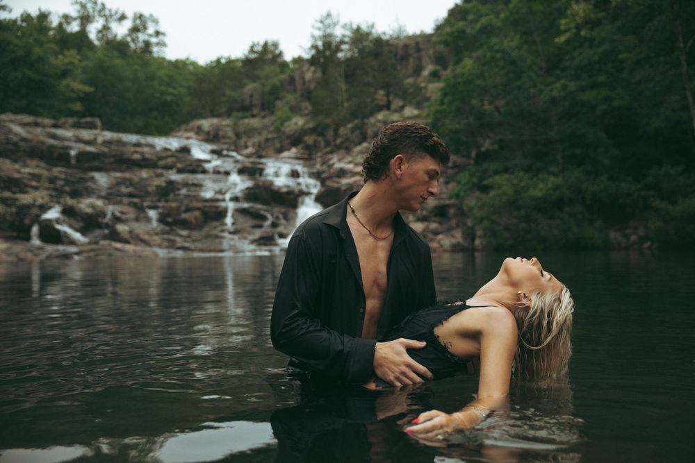 Couple embracing in a natural rock pool with a waterfall and lush greenery in the background.