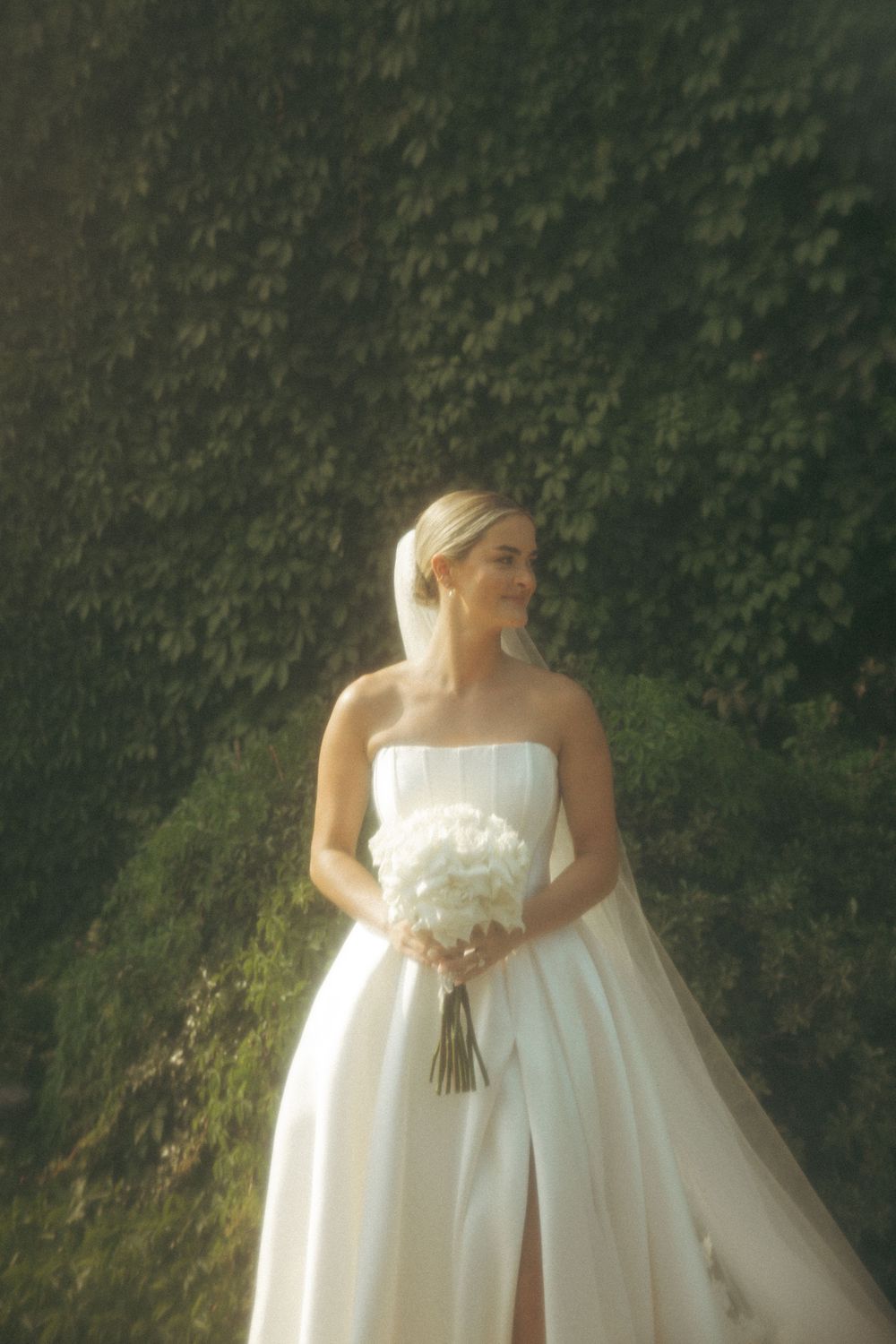 A bride in a flowing white empire waist wedding gown stands against a lush green ivy-covered background.