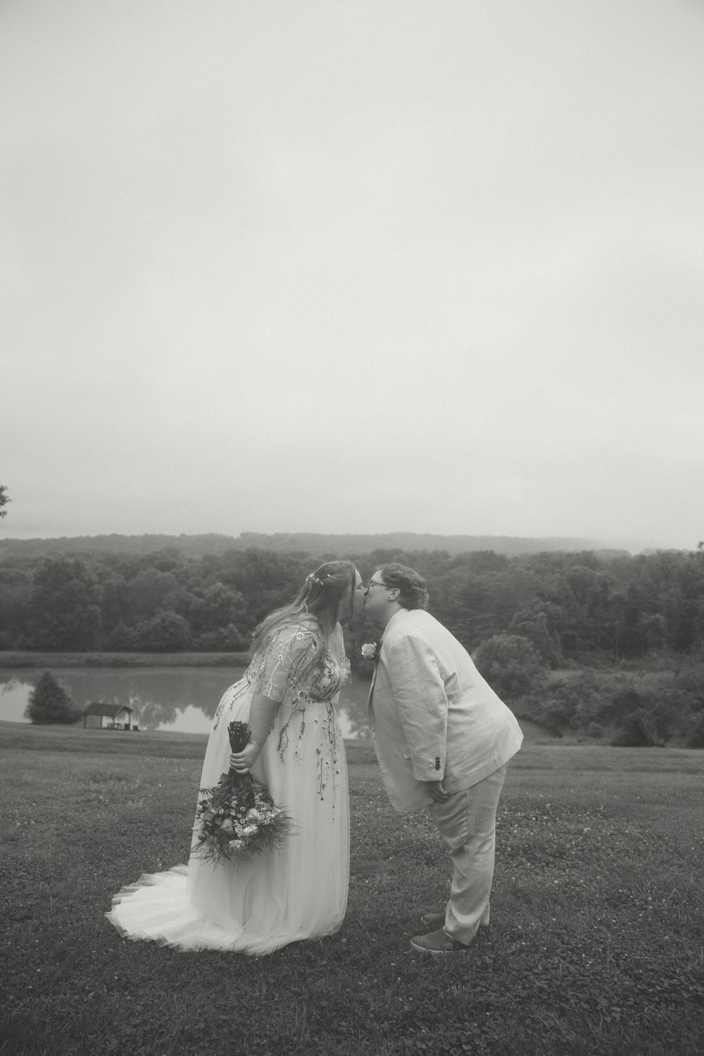 Black and white photo of a bride and groom sharing a kiss outdoors on their wedding day with a scenic landscape behind them.