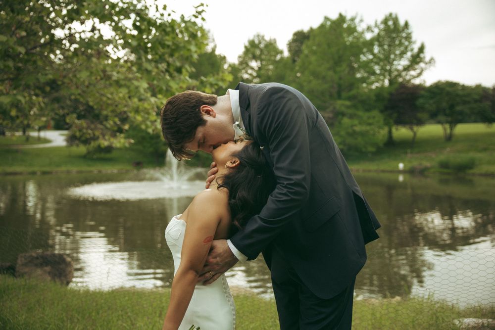 A bride and groom share a romantic kiss near a serene pond surrounded by lush green trees on their wedding day.