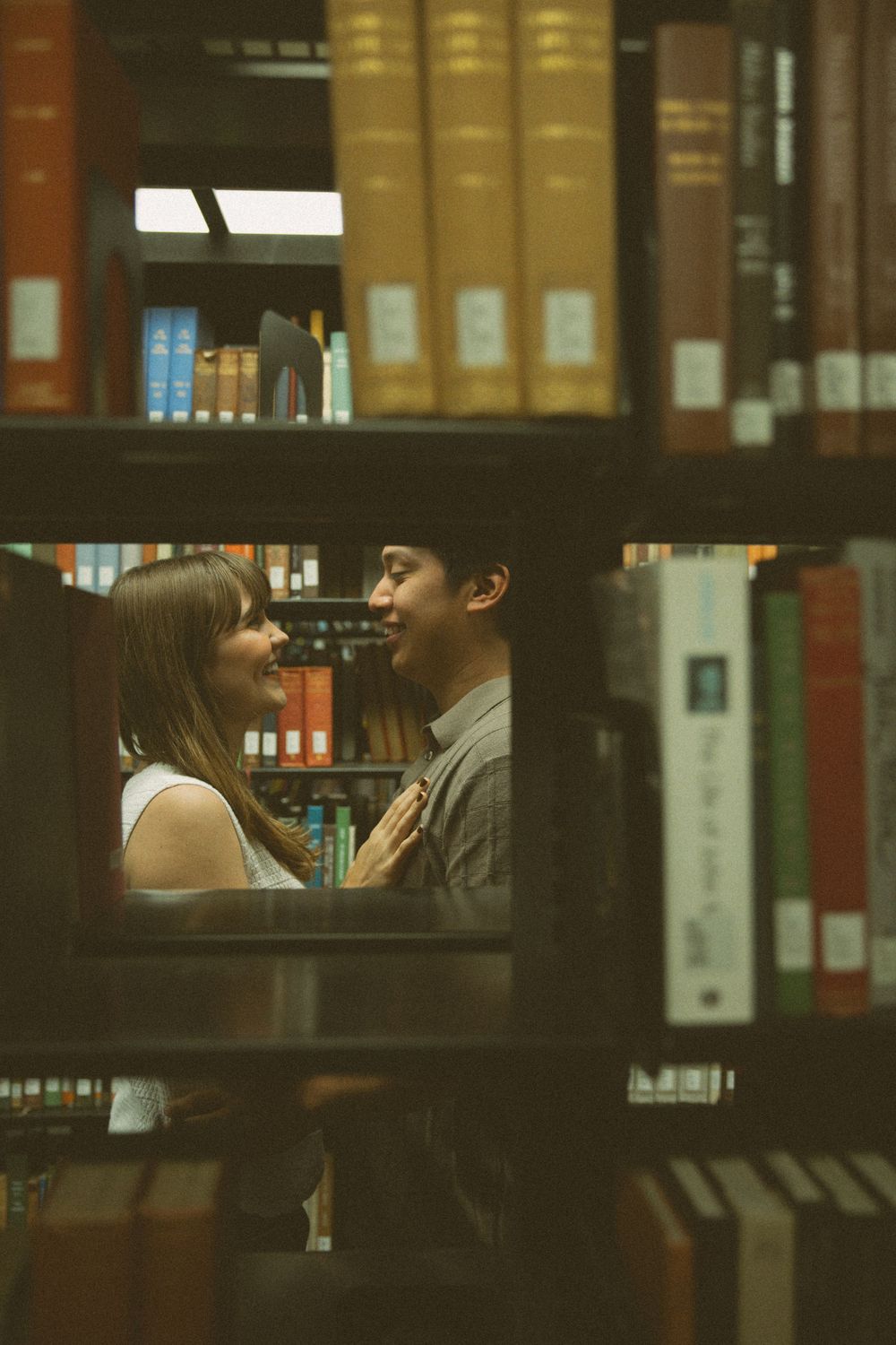 A couple shares an intimate moment between library bookshelves in moody, warm lighting.