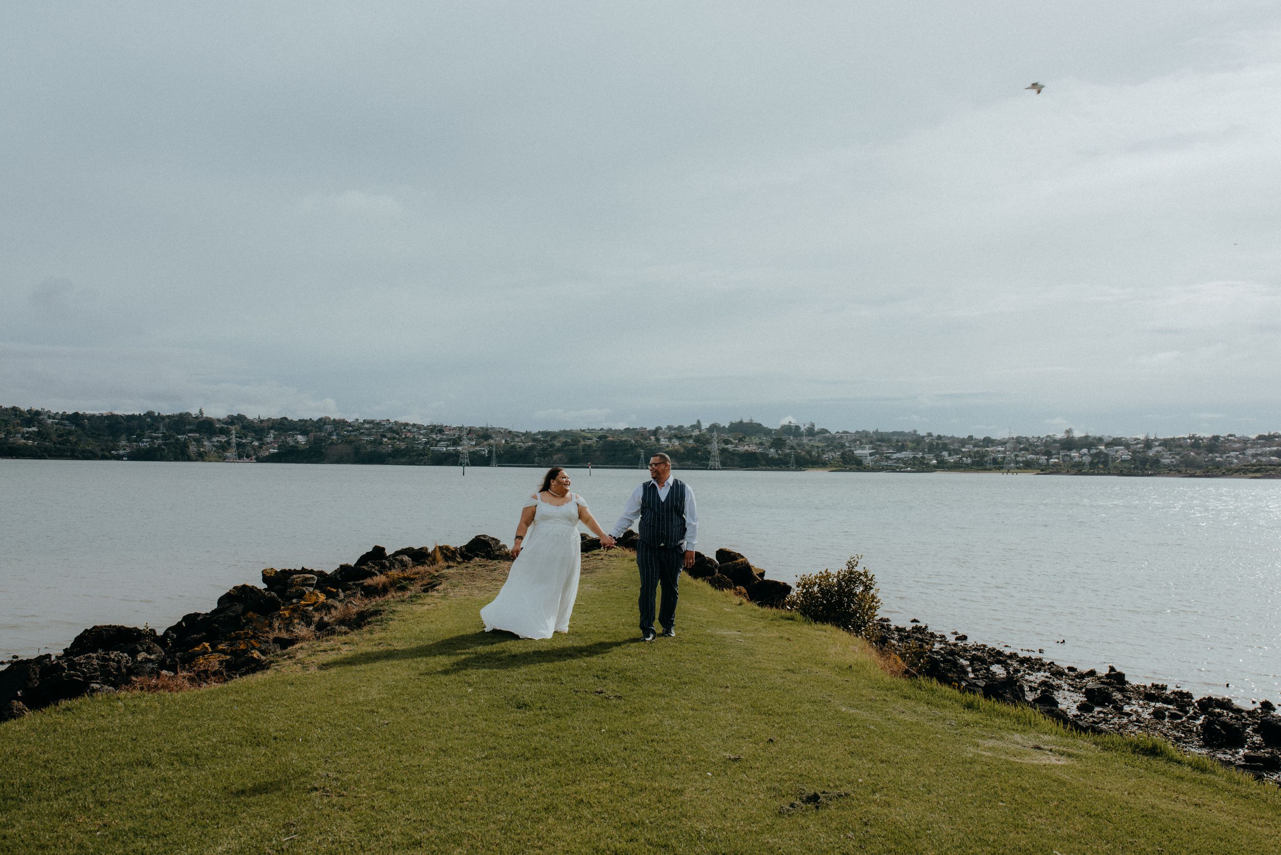 Bride and groom holding hands on grassy cliff overlooking scenic harbor at outdoor wedding.