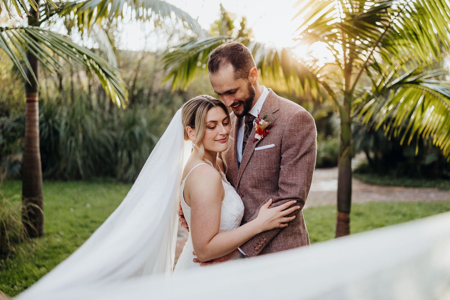 Bride and groom embrace outdoors surrounded by palm trees, bride wearing a flowing veil in a romantic tropical wedding setting.