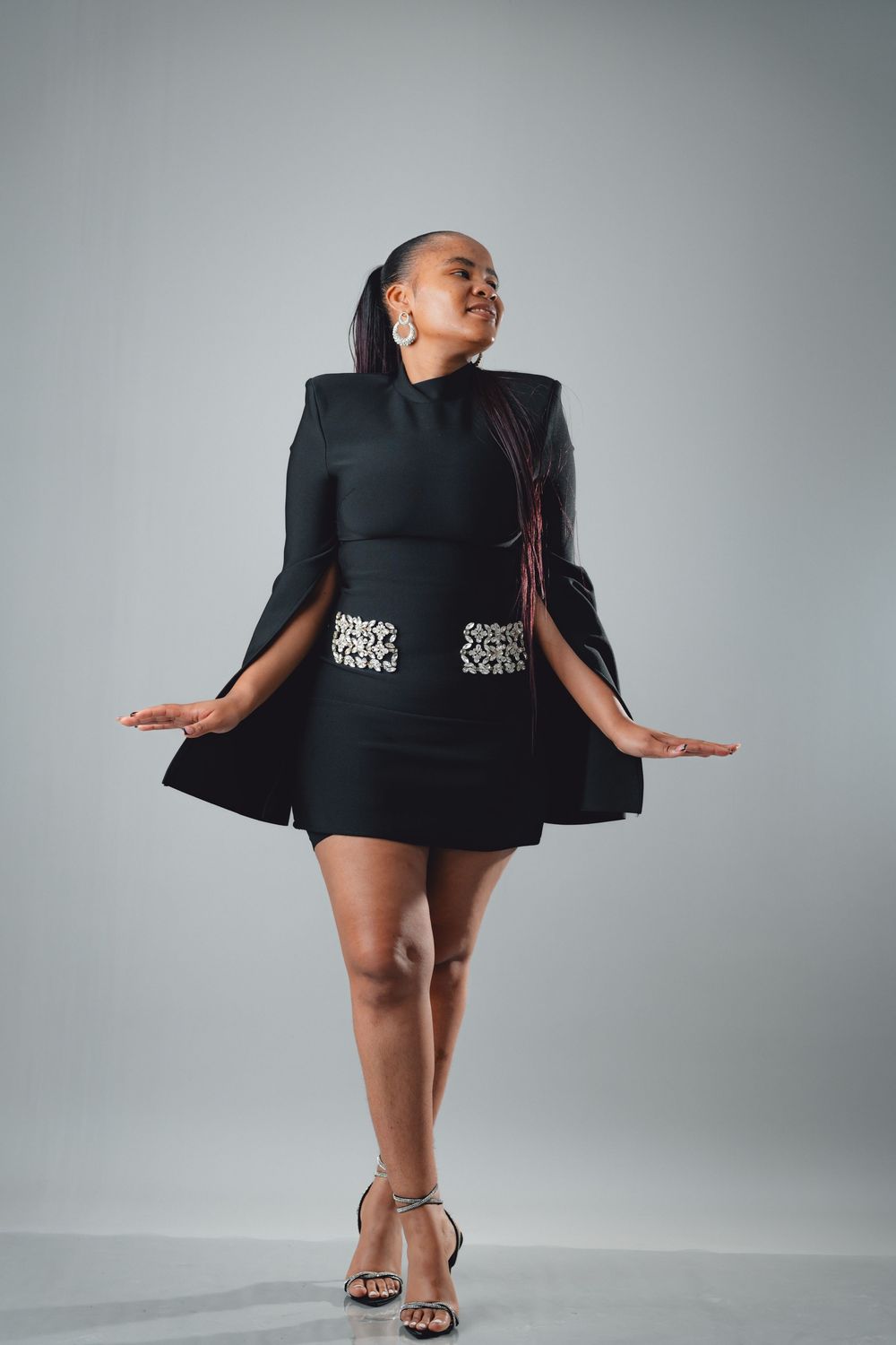Fashion model wearing black dress with structured shoulders and silver sandals poses against grey studio backdrop.