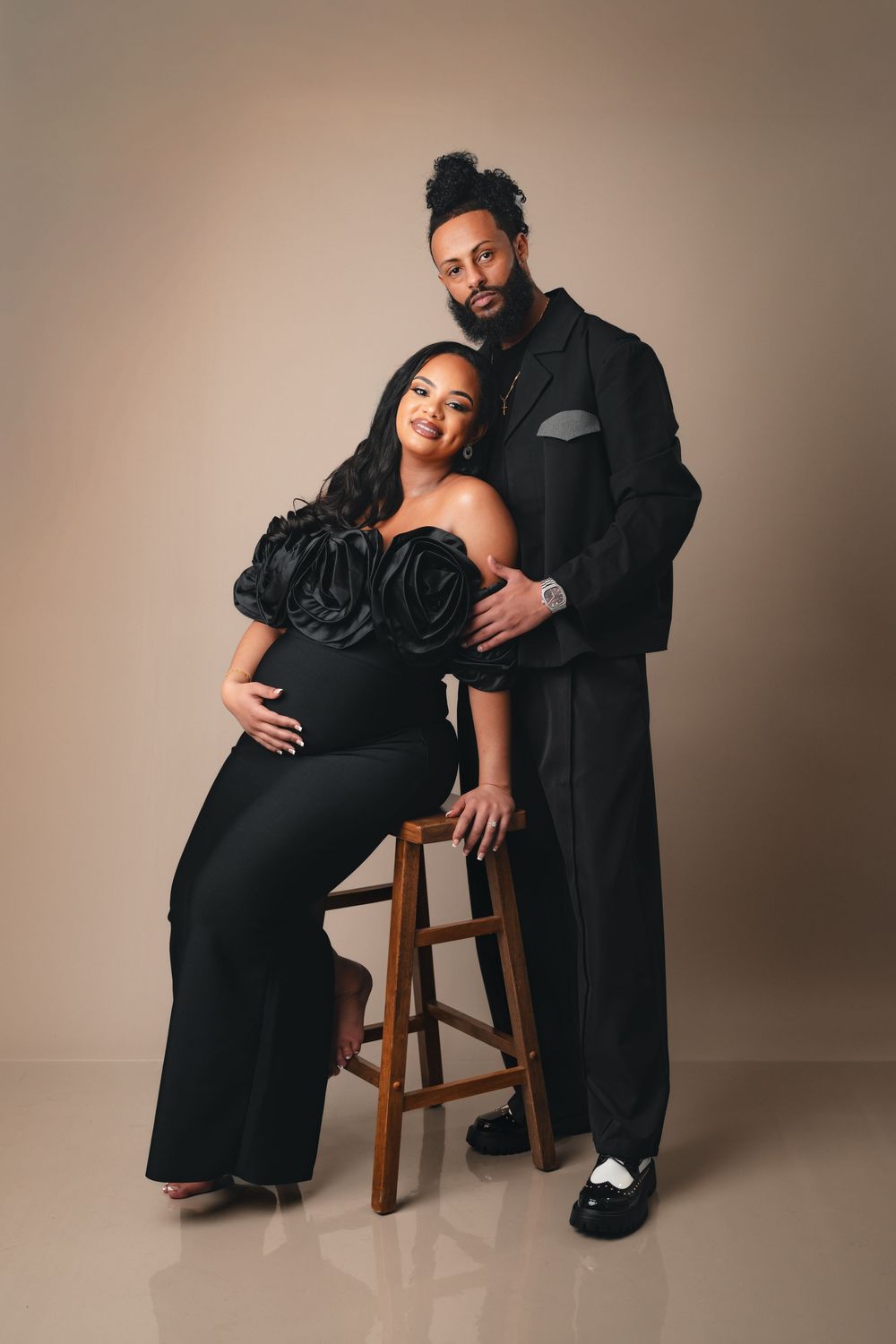 A maternity photoshoot series showing a couple in black attire posing intimately with a wooden stool against a neutral backdrop.