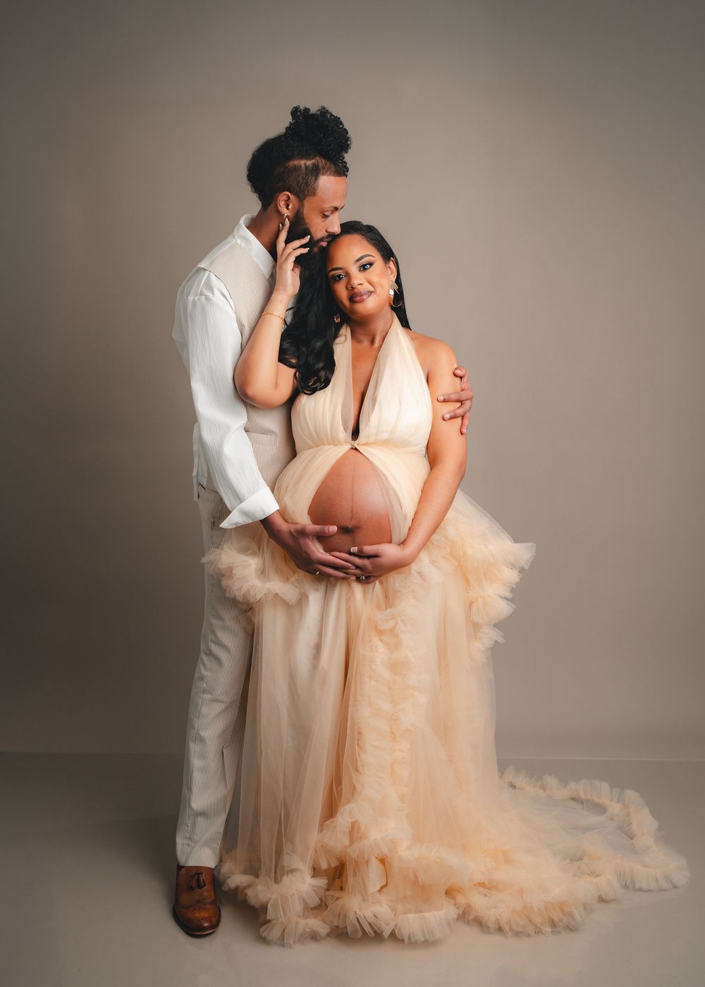Romantic maternity photo of couple in white and peach formal wear embracing against neutral studio backdrop.