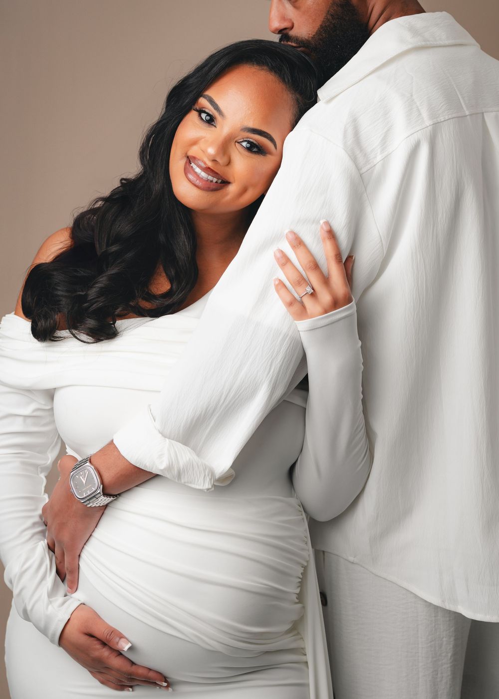Couple in white outfits embracing during a maternity photoshoot against neutral background.