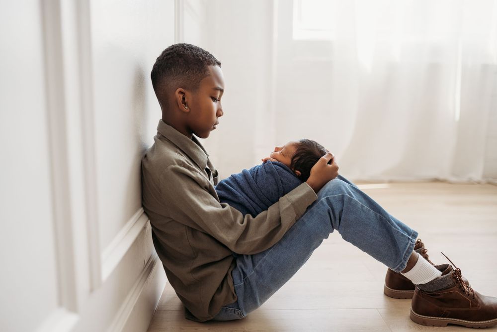 boy sitting on floor leaning on wall holding his newborn baby brother who is wrapped in a navy blue swaddle.