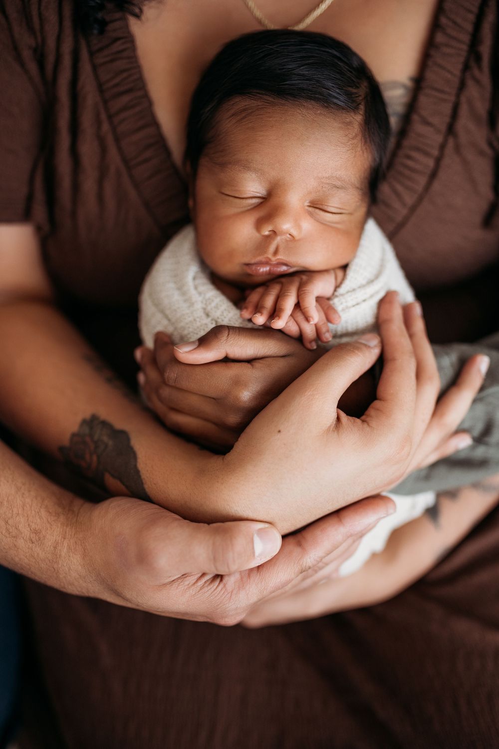 Newborn in ivory swaddle wrapped by 3 sets of hands laying on mothers chest.