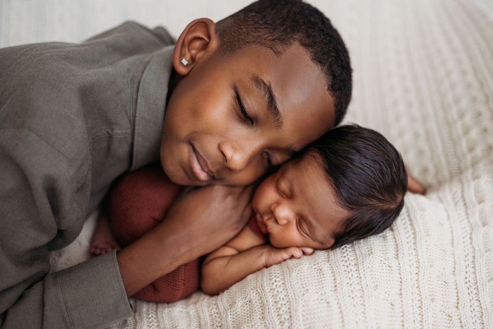 newborn laying on ivory blanket while big brother rests his head on him smiling.