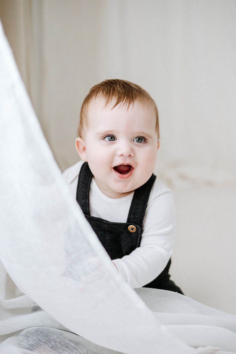 A baby wearing a black and white outfit peeks through white fabric with a delighted expression.