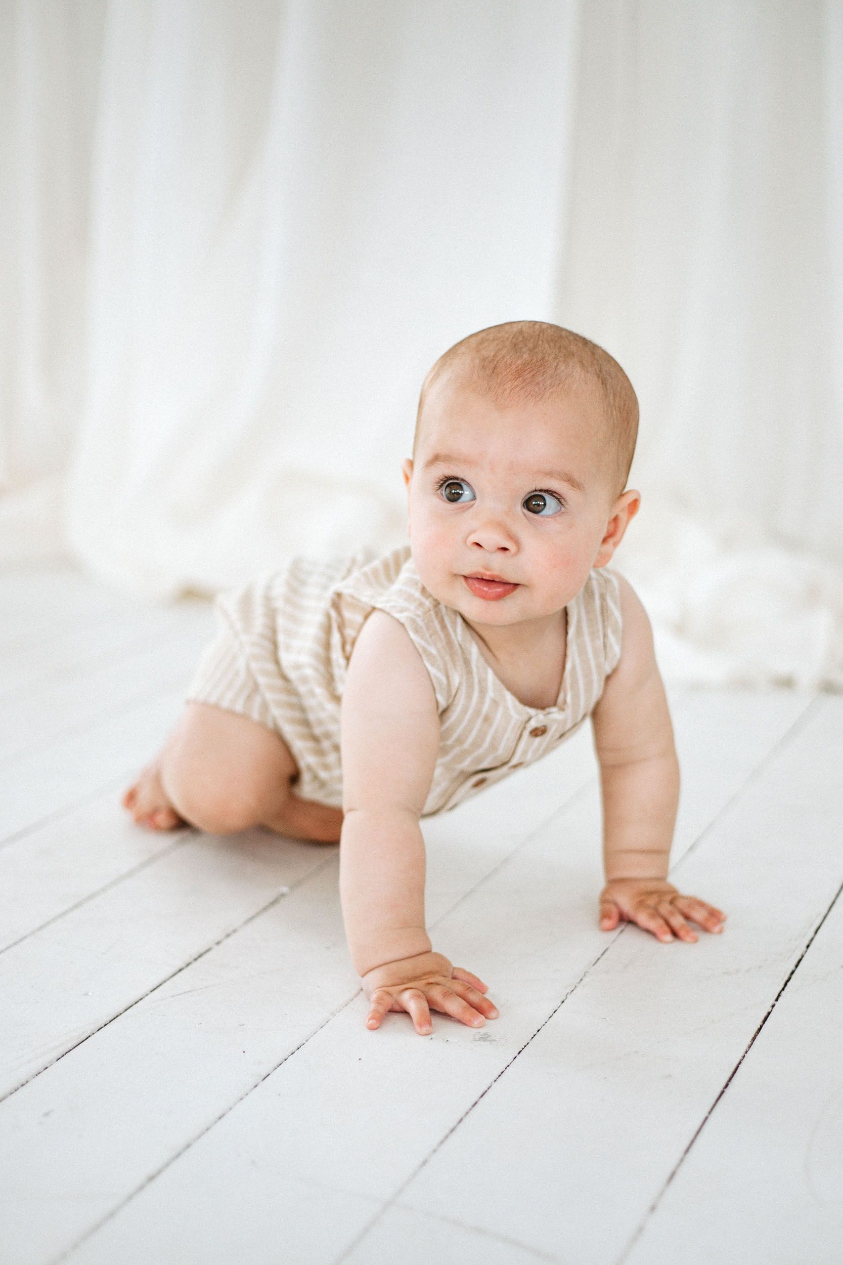 A curious baby in a striped onesie crawls on a white wooden floor against a soft white backdrop.