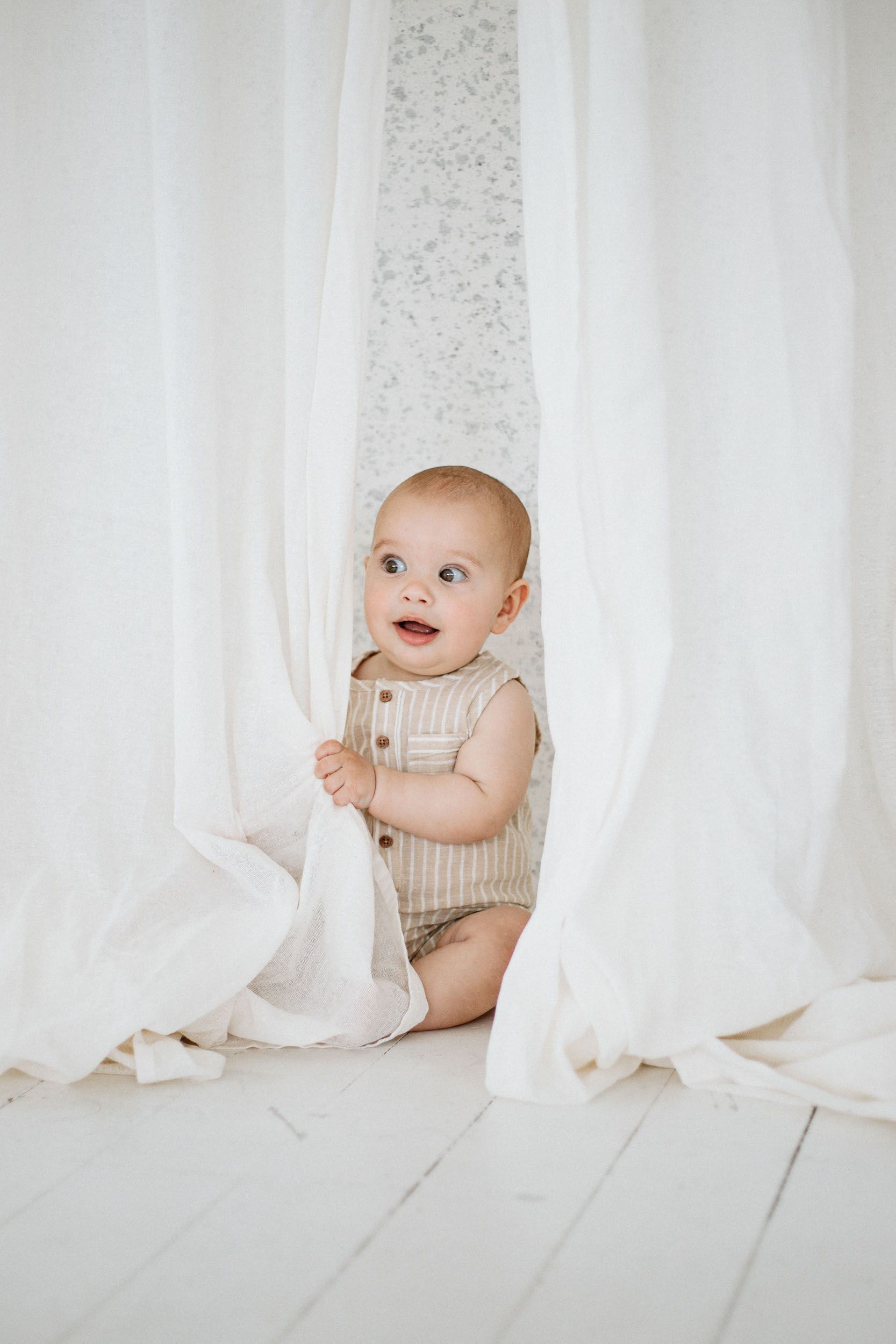 Adorable baby in a striped romper peeks through white curtains on a white wooden floor.