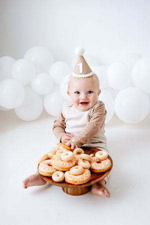 A series of photos showing a baby in white sitting with a wooden platter of frosted donuts against a light background.