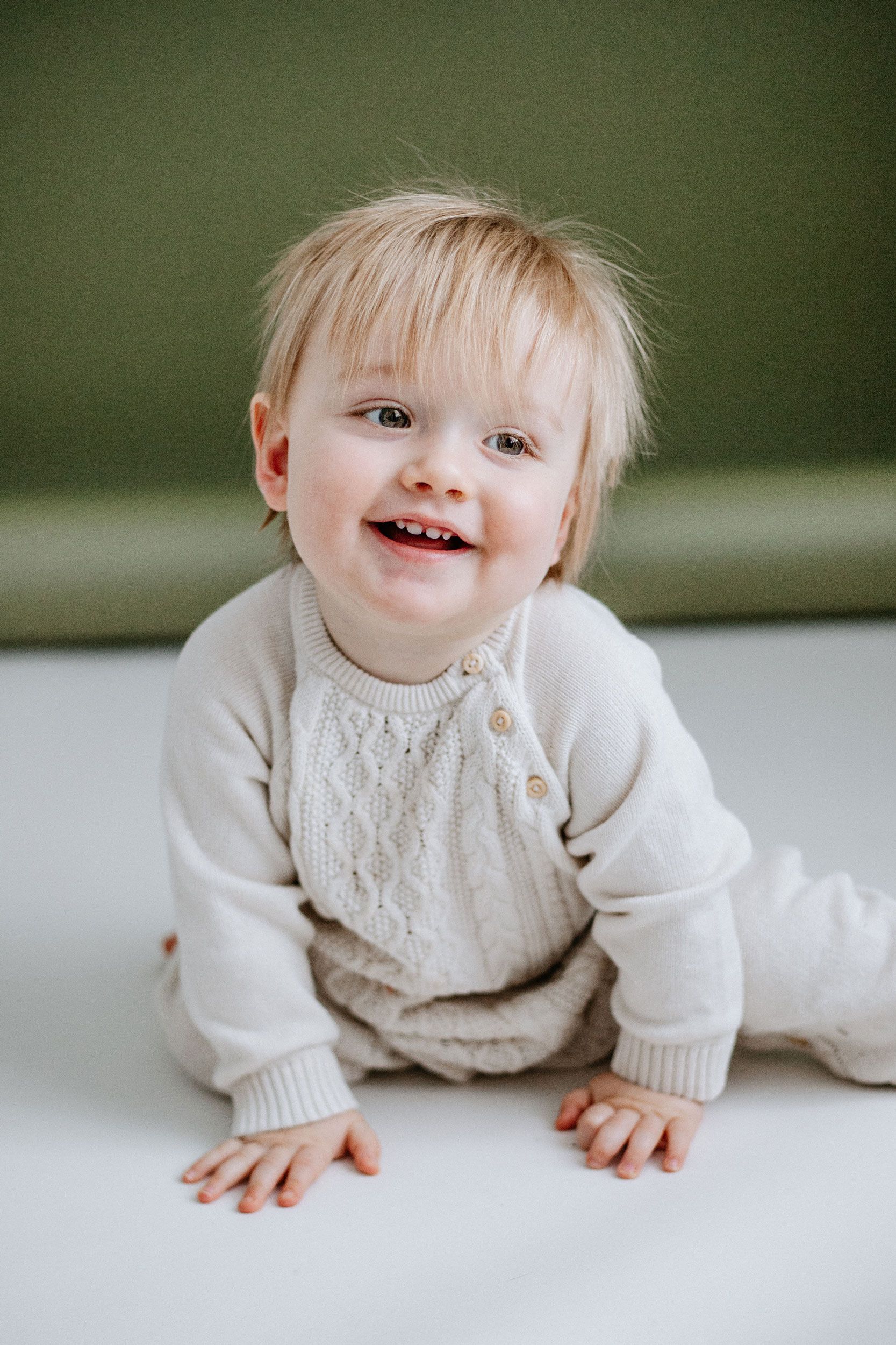 Smiling baby in a cream knit romper sitting on a white surface with a green background.