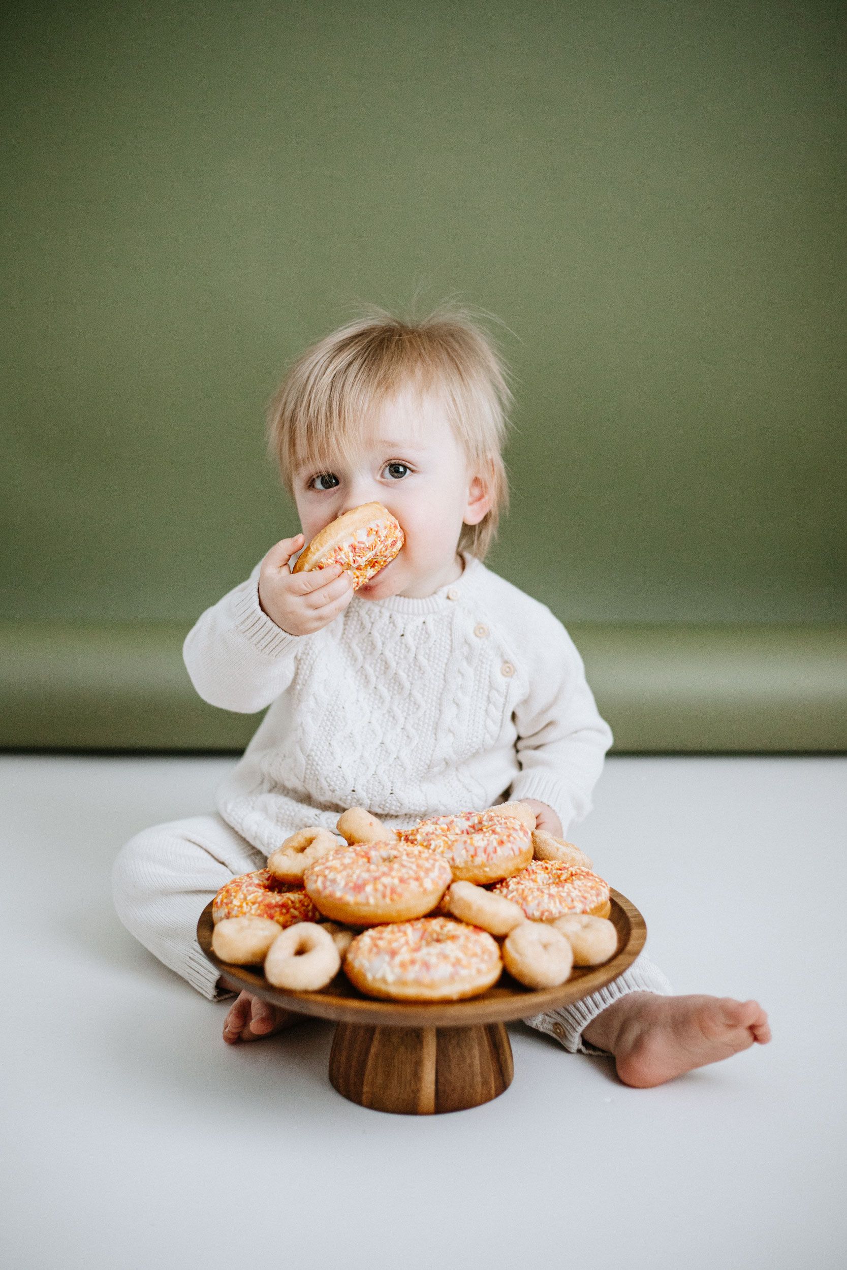 Toddler in white outfit eating an orange sprinkled donut from a wooden cake stand filled with donuts.