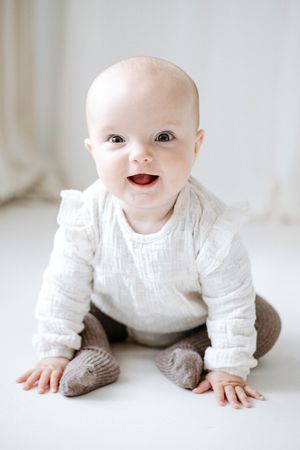 A baby in white clothing crawls and smiles on a light-colored floor.