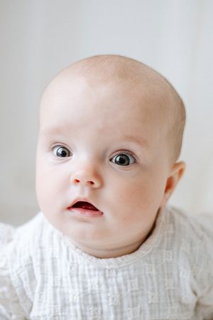 6 month old baby in a white long sleeved top staring inquisitively at the camera.