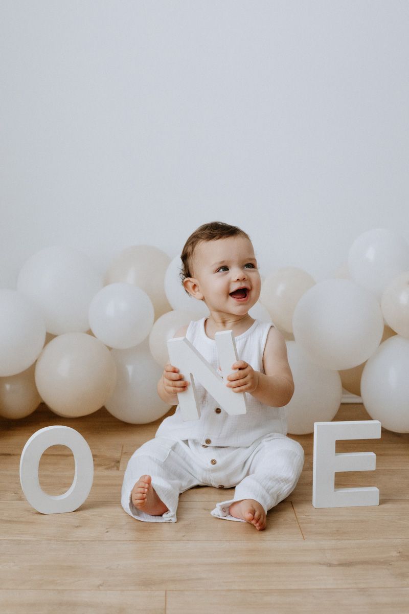 Baby boy in white outfit sits smiling among white balloons and ONE letters for first birthday photoshoot in Mansfield
