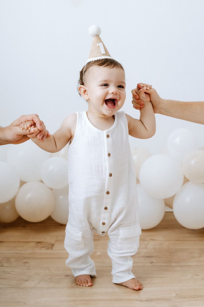 A joyful baby in a white romper and birthday hat stands barefoot, held by adult hands, with white balloons in the background.