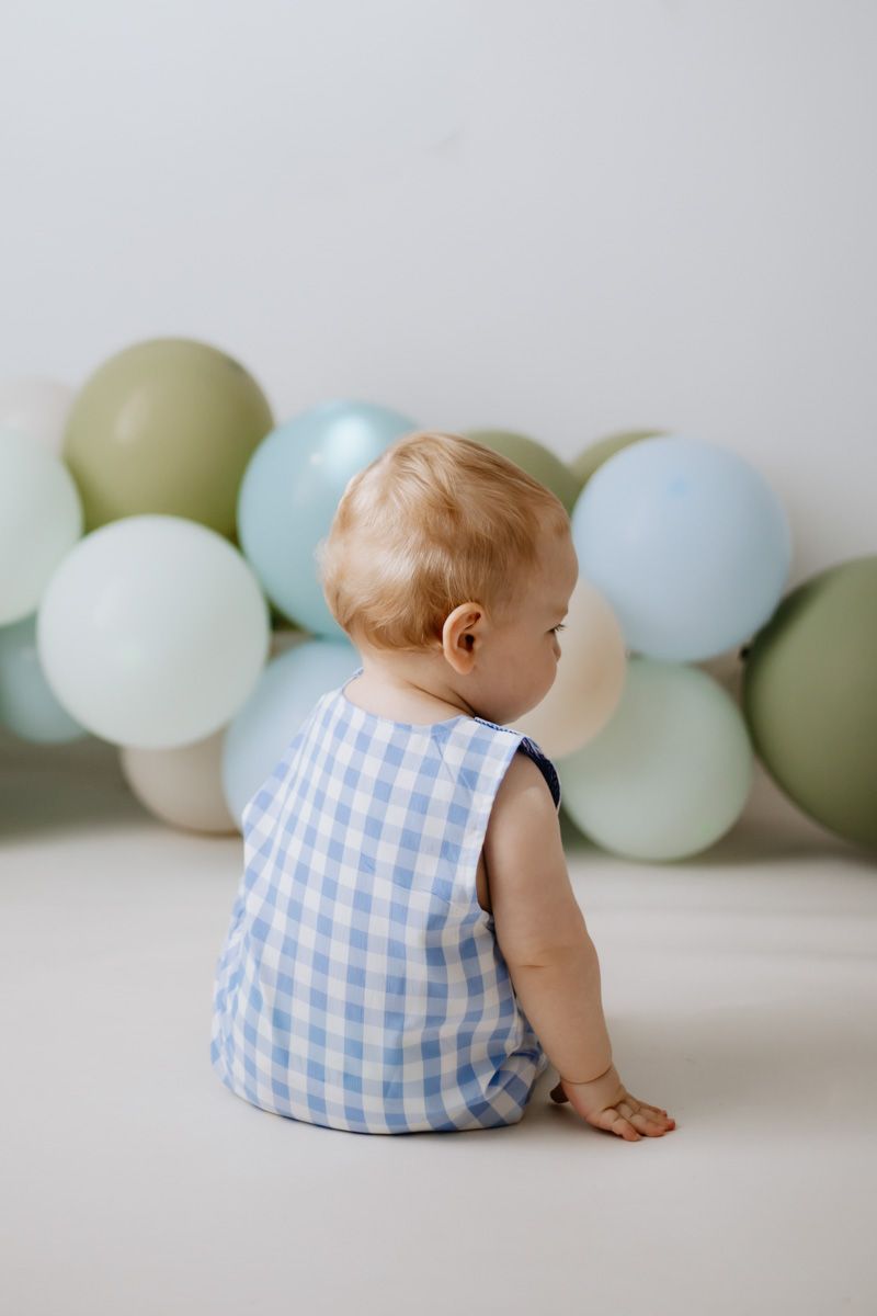 A baby in a blue gingham outfit sits on the floor surrounded by pastel green and blue balloons.