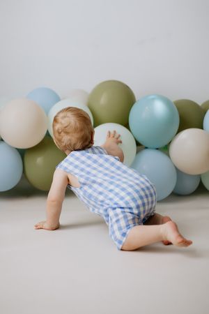 Baby in blue gingham romper crawling toward colorful pastel balloons on white background.