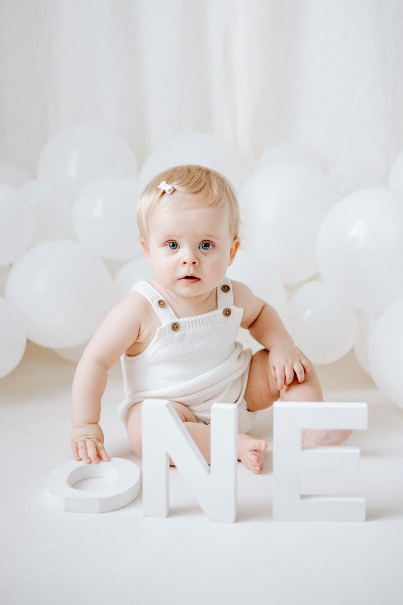 A baby in white overalls sits on a white backdrop with linen curtain effect next to decorative letter blocks spelling ONE.
