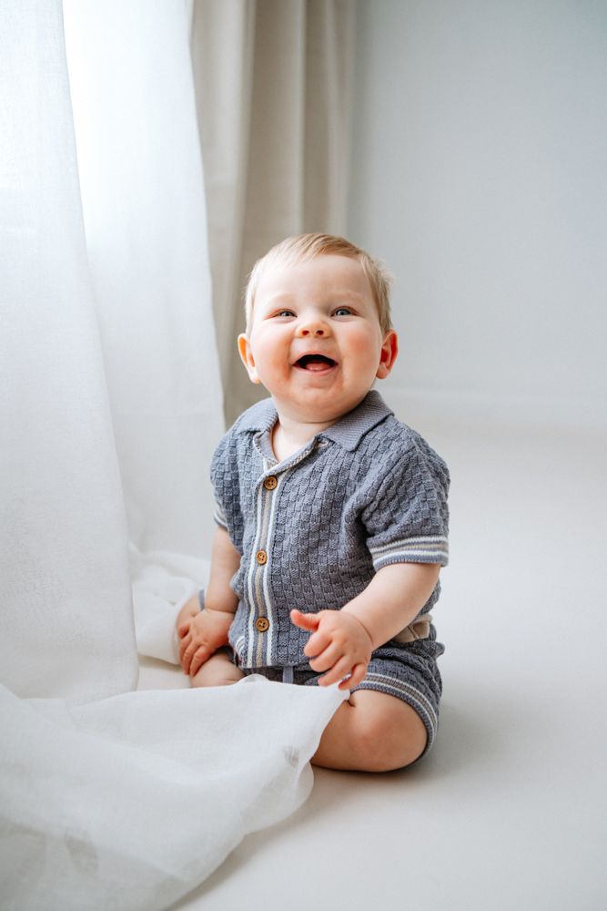 Smiling baby wearing grey blue cardigan sits on white backdrop in series of lifestyle portraits.
