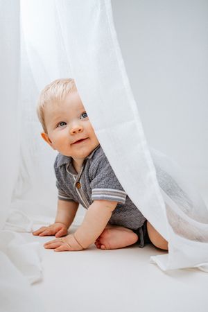 Sweet baby in blue knitted outfit plays and smiles while crawling under flowing white curtains in bright natural light.