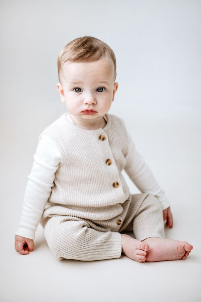 A baby wearing a cosy cream-colored knit sweater with buttons sits on a white background in various poses.