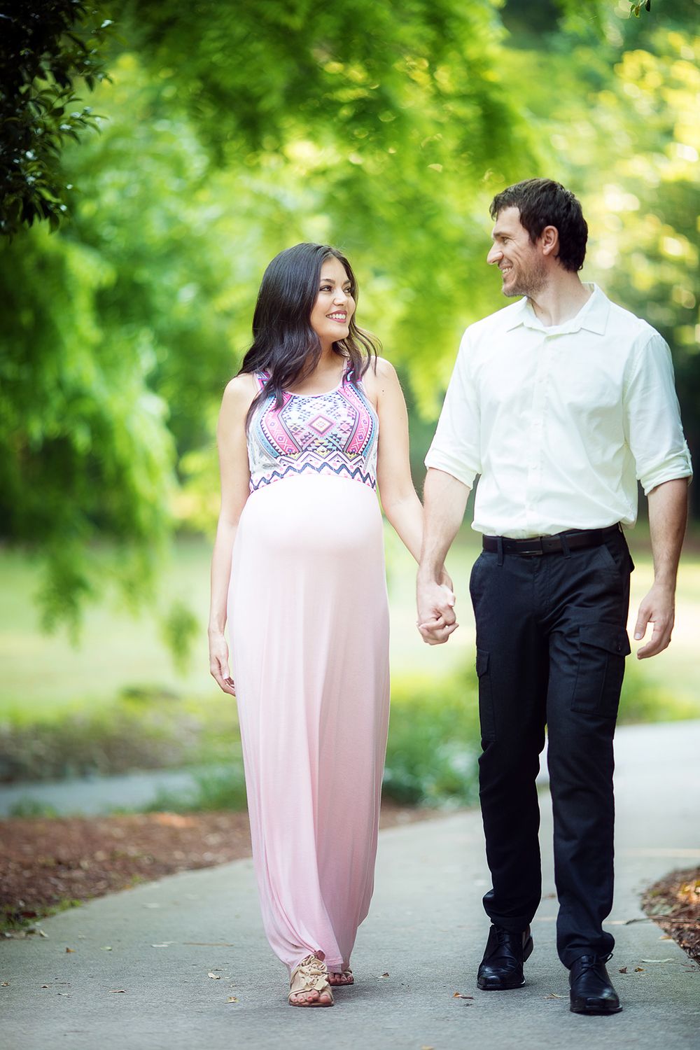A couple in pink and white attire walks together on a wooded path during an engagement photo session.