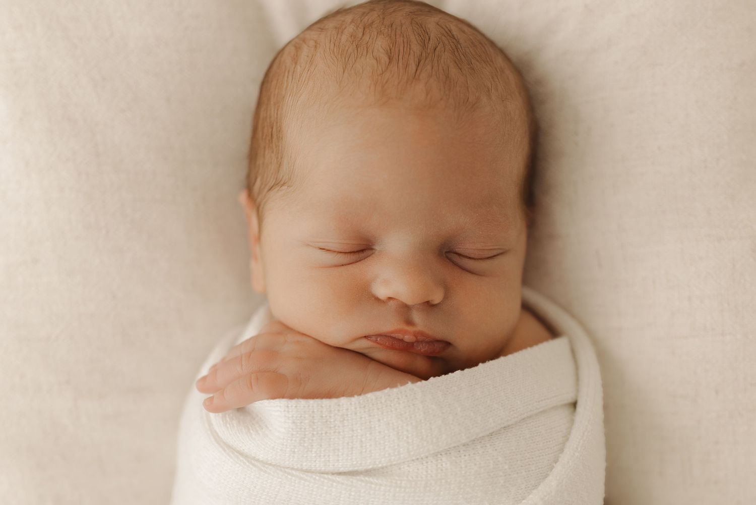 A sleeping newborn baby wearing a white bow headband rests peacefully in gentle hands.