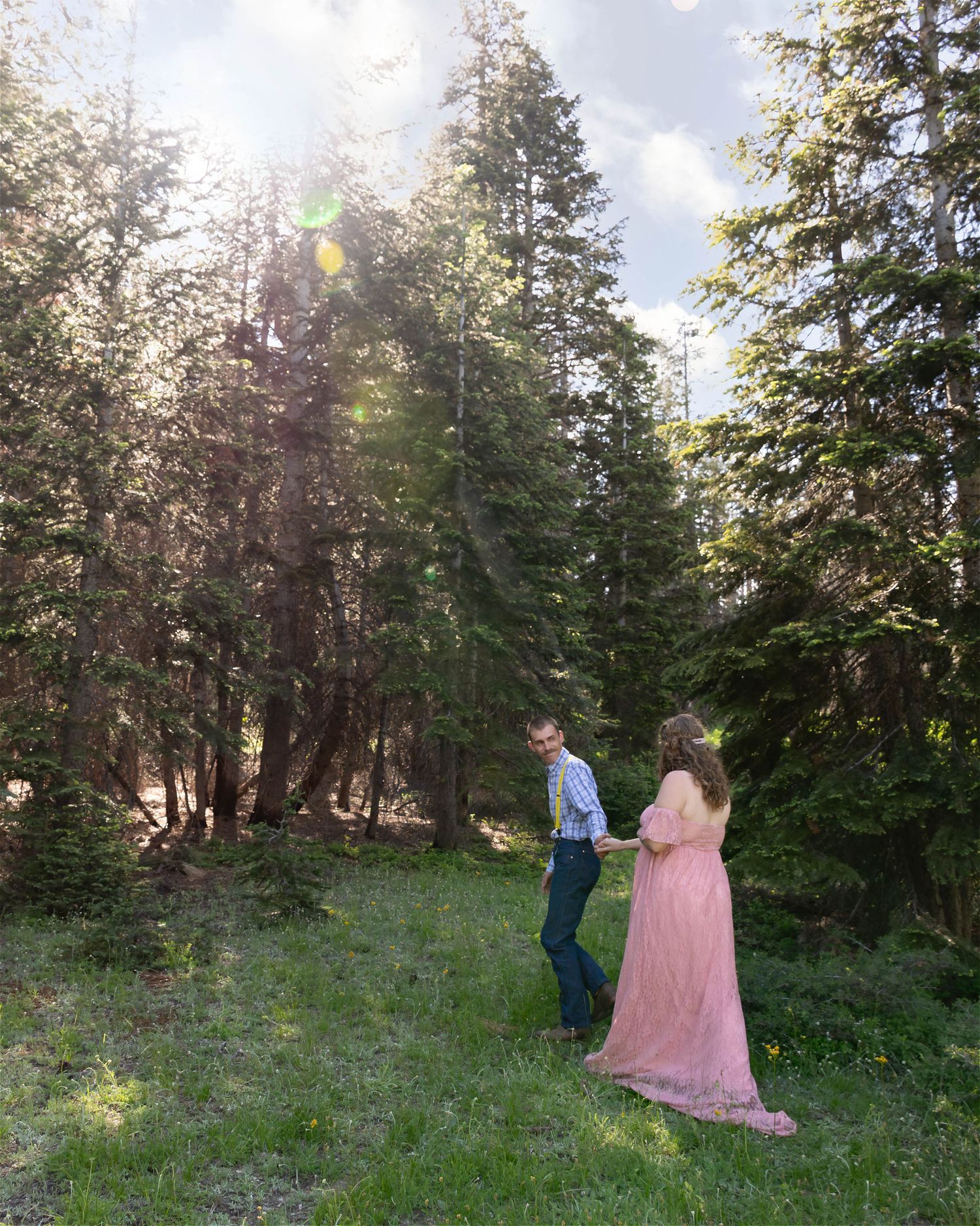 Couple stands in a sunlit forest clearing with tall evergreen trees.