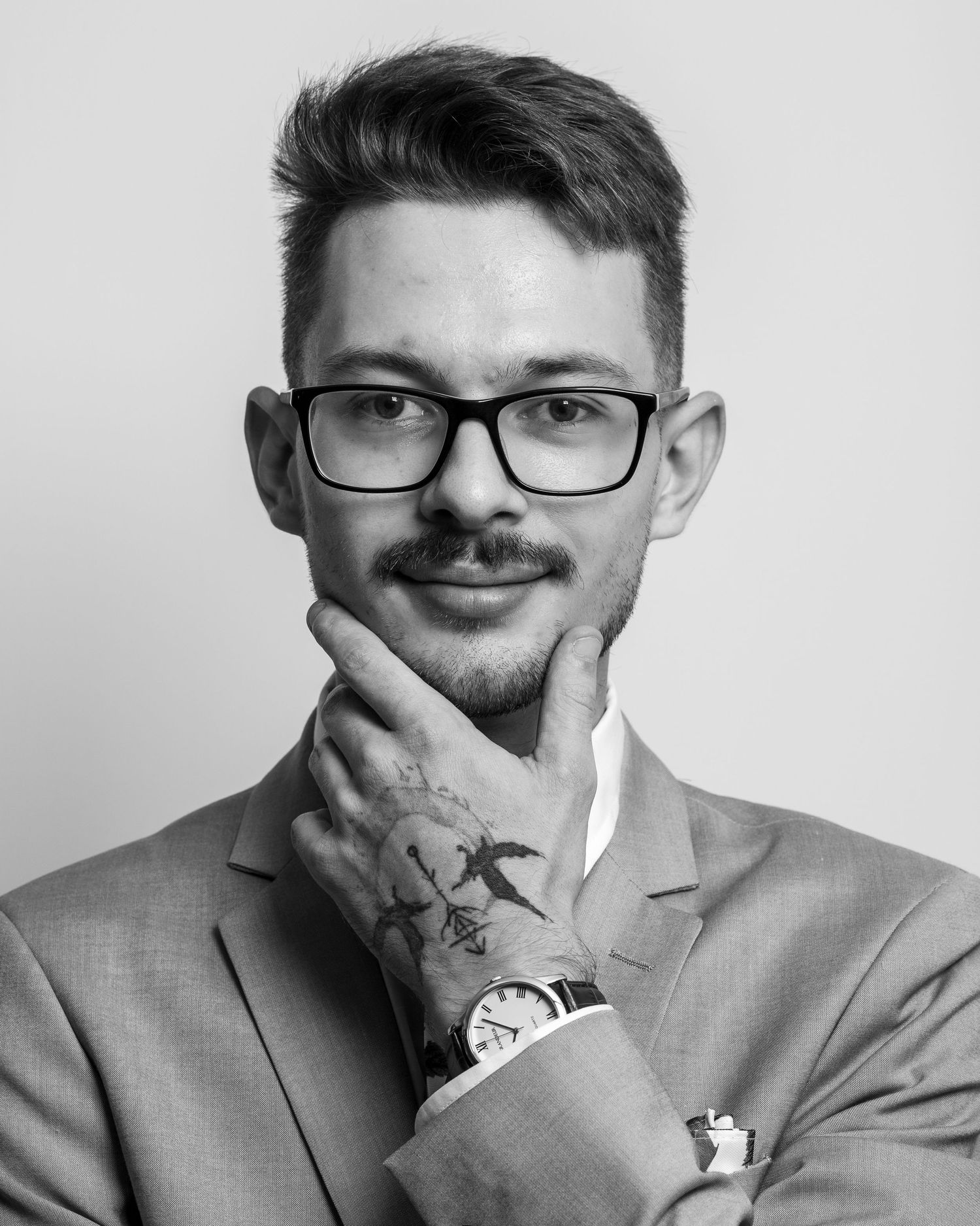 Professional headshot of a person in glasses and grey suit with hand tattoo posing against white background.