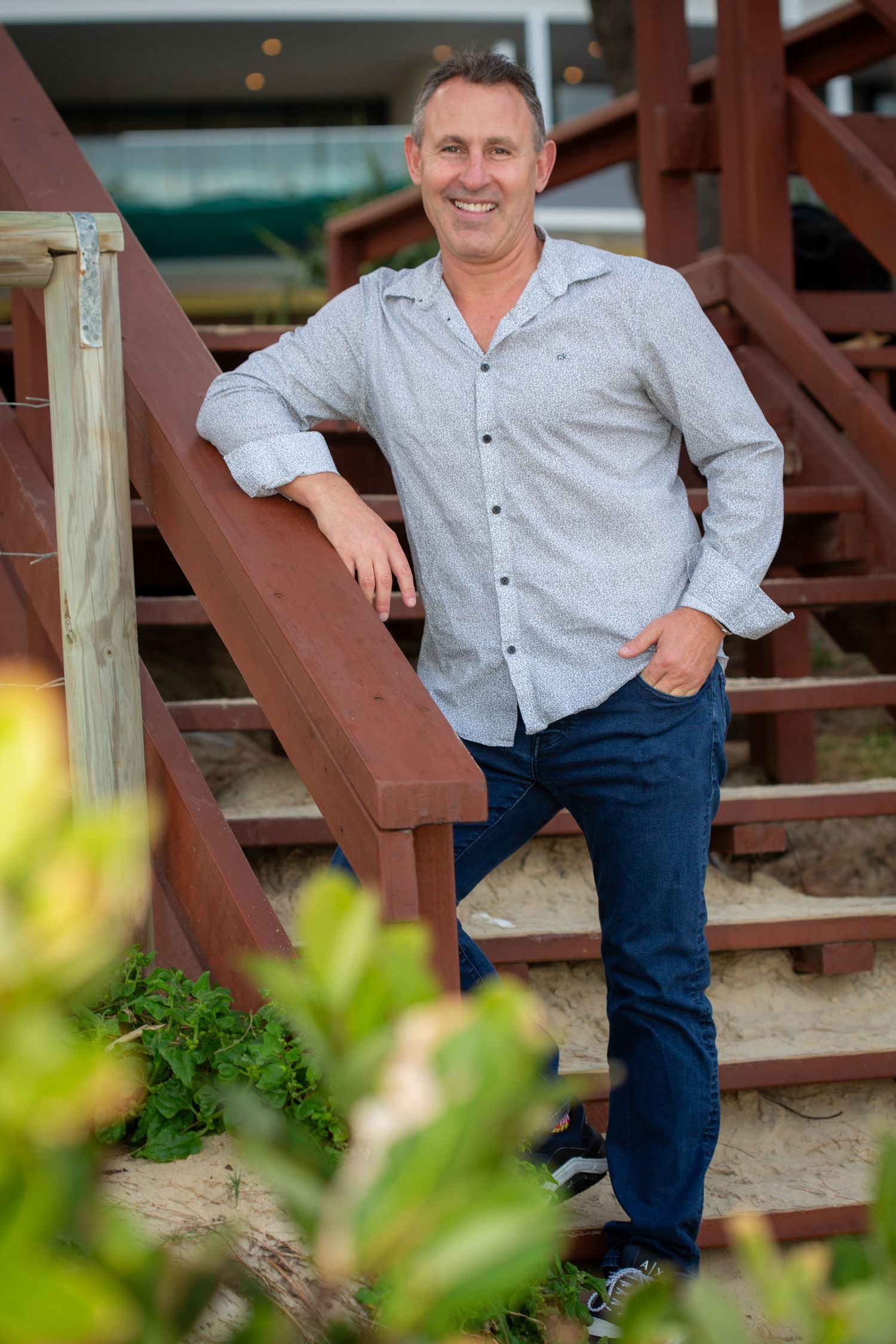 A casual portrait of a person in a grey button-down shirt posing on wooden stairs outdoors.