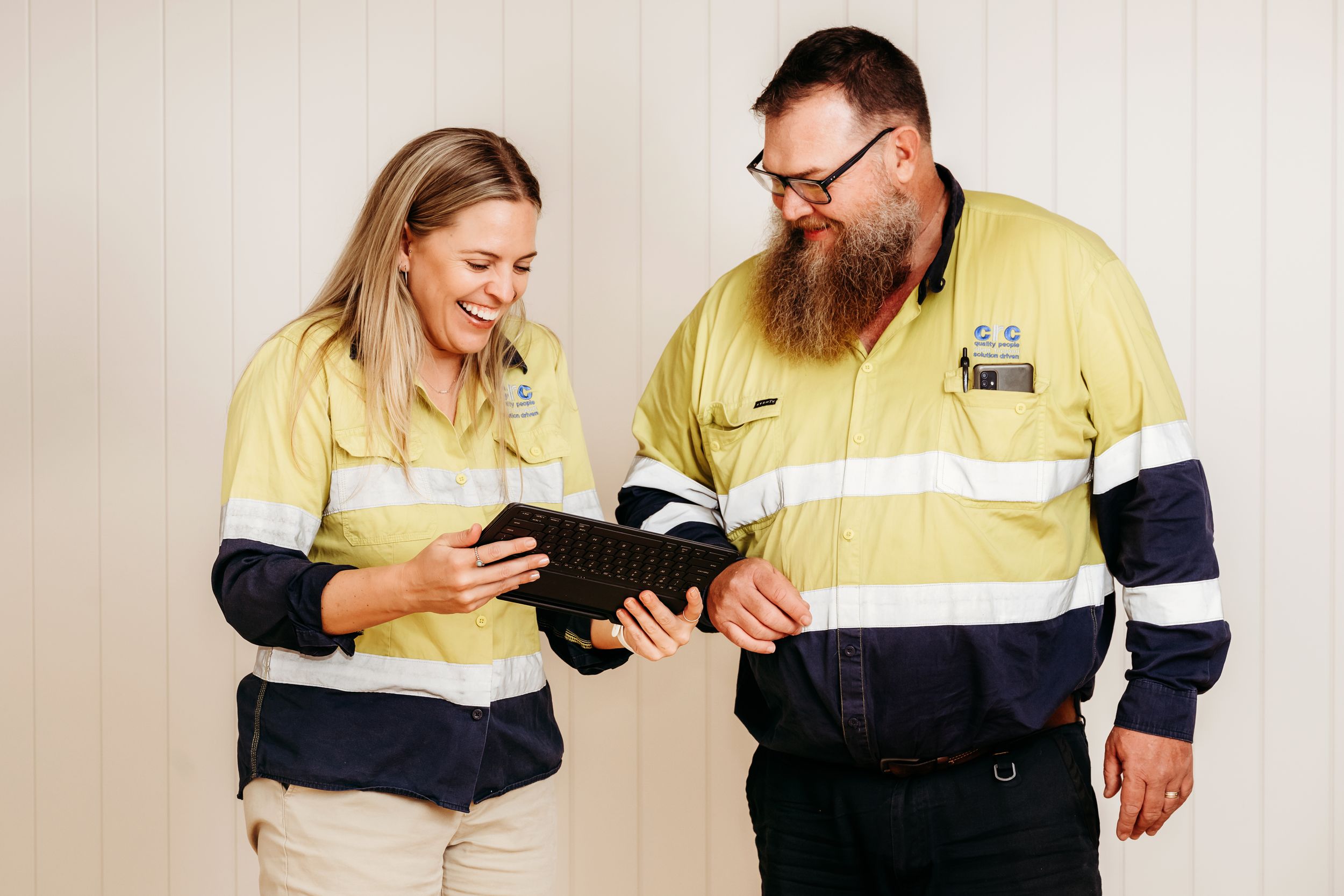Two workers in hi-vis yellow and navy uniforms examine paperwork while standing together in a bright setting.