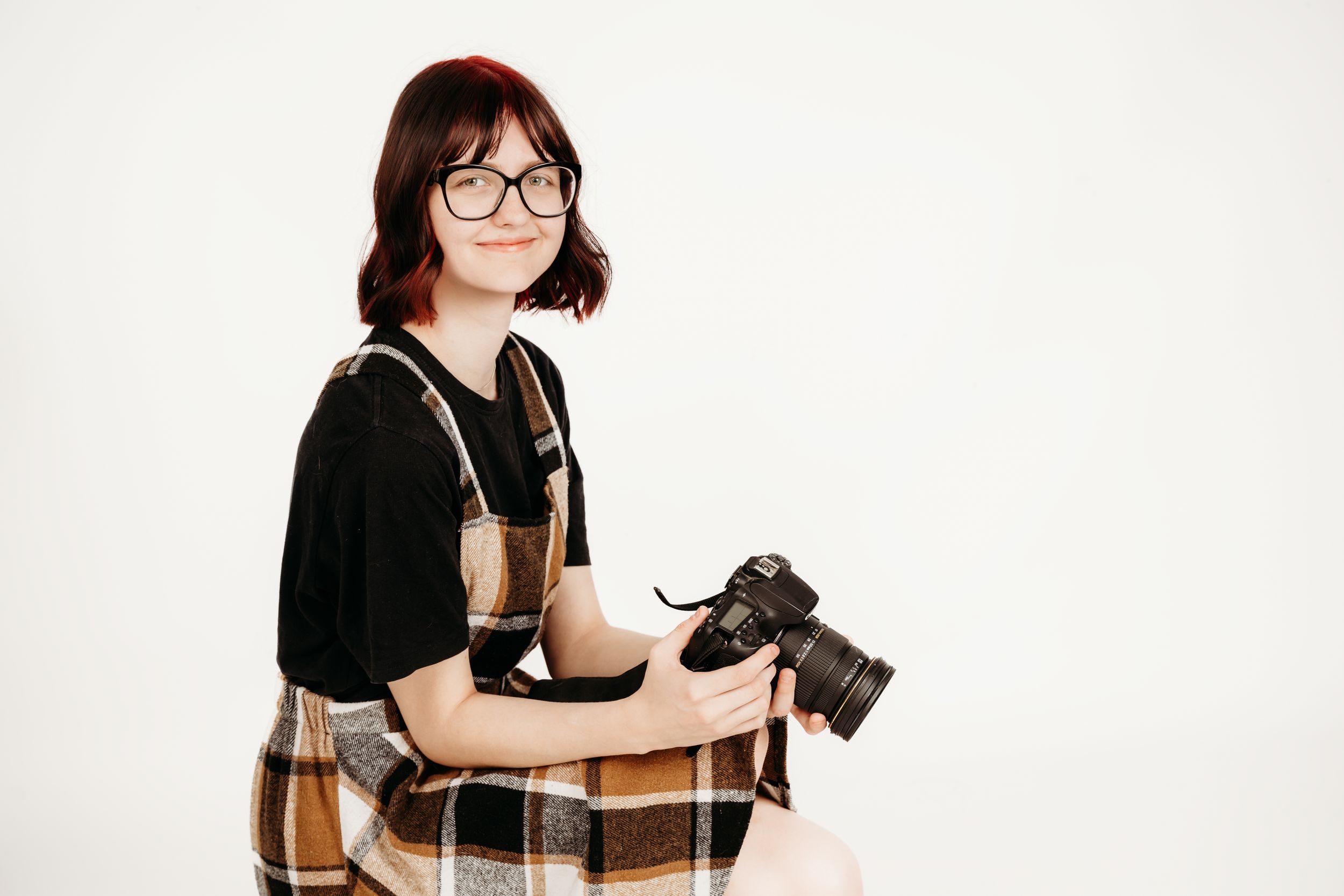 A photographer wearing glasses and plaid dress holds a professional camera while sitting against white background.
