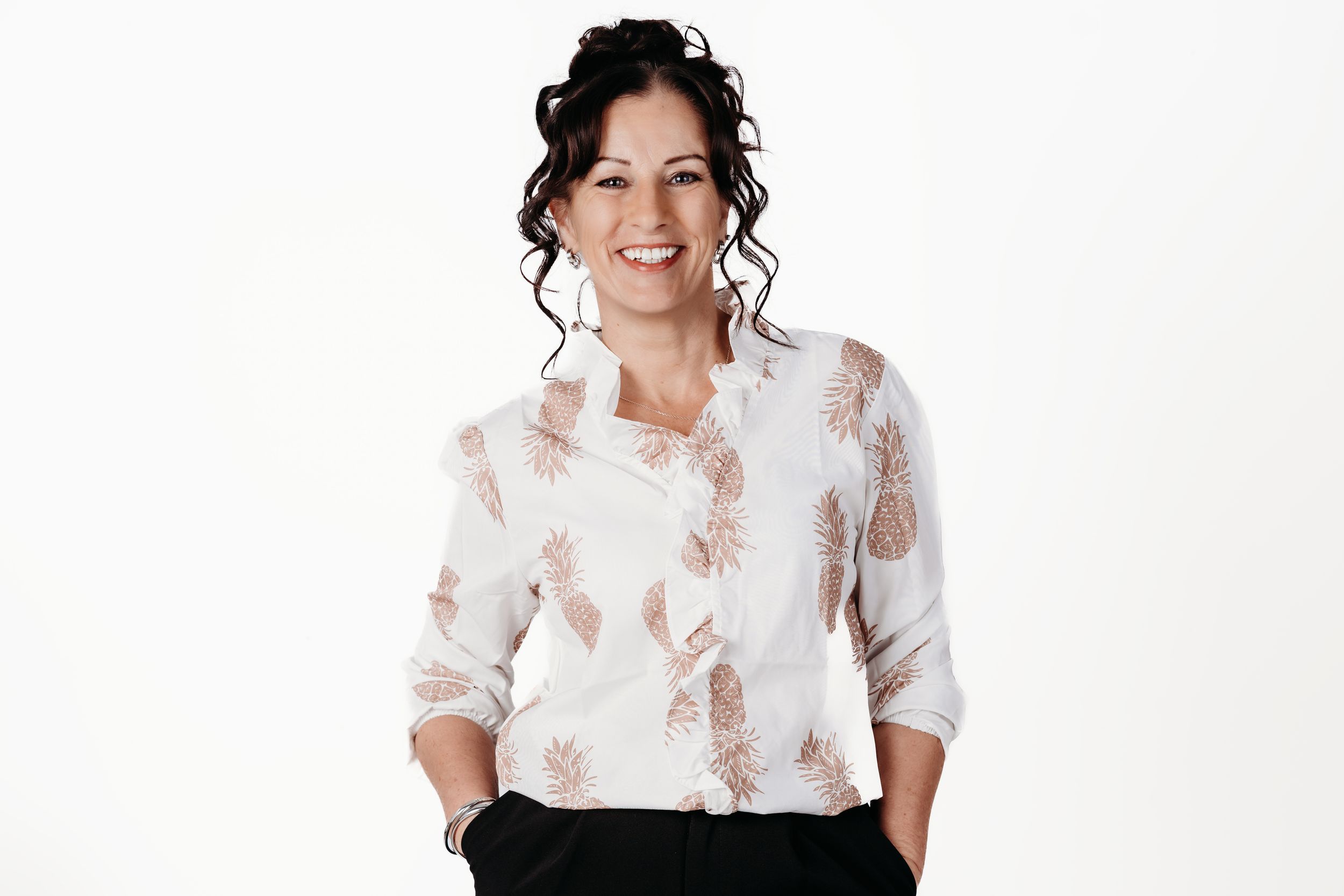 Smiling woman in white floral blouse with hands in pockets against white background.