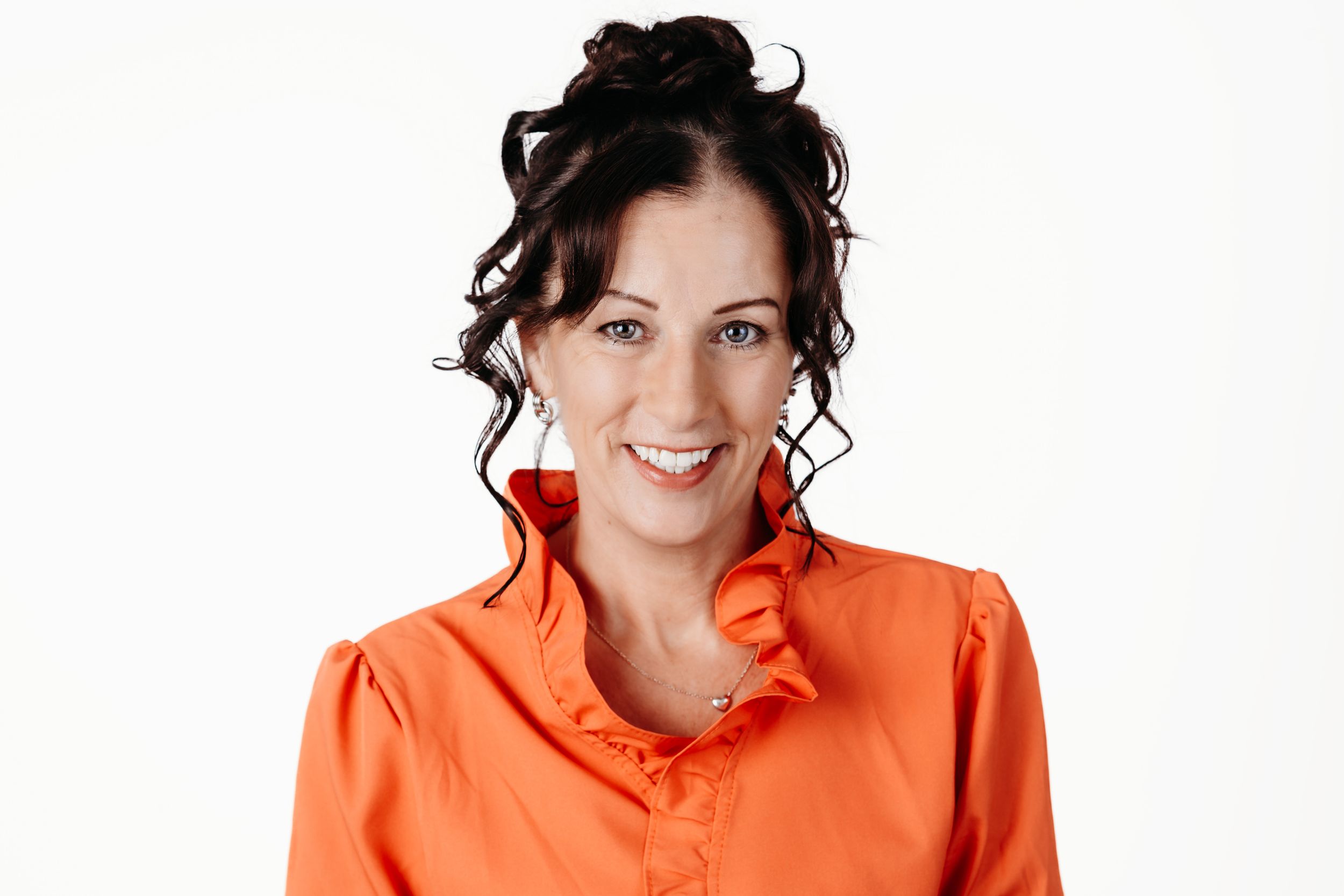Smiling woman with dark curly updo hair wearing a bright orange ruffled blouse against a white background.