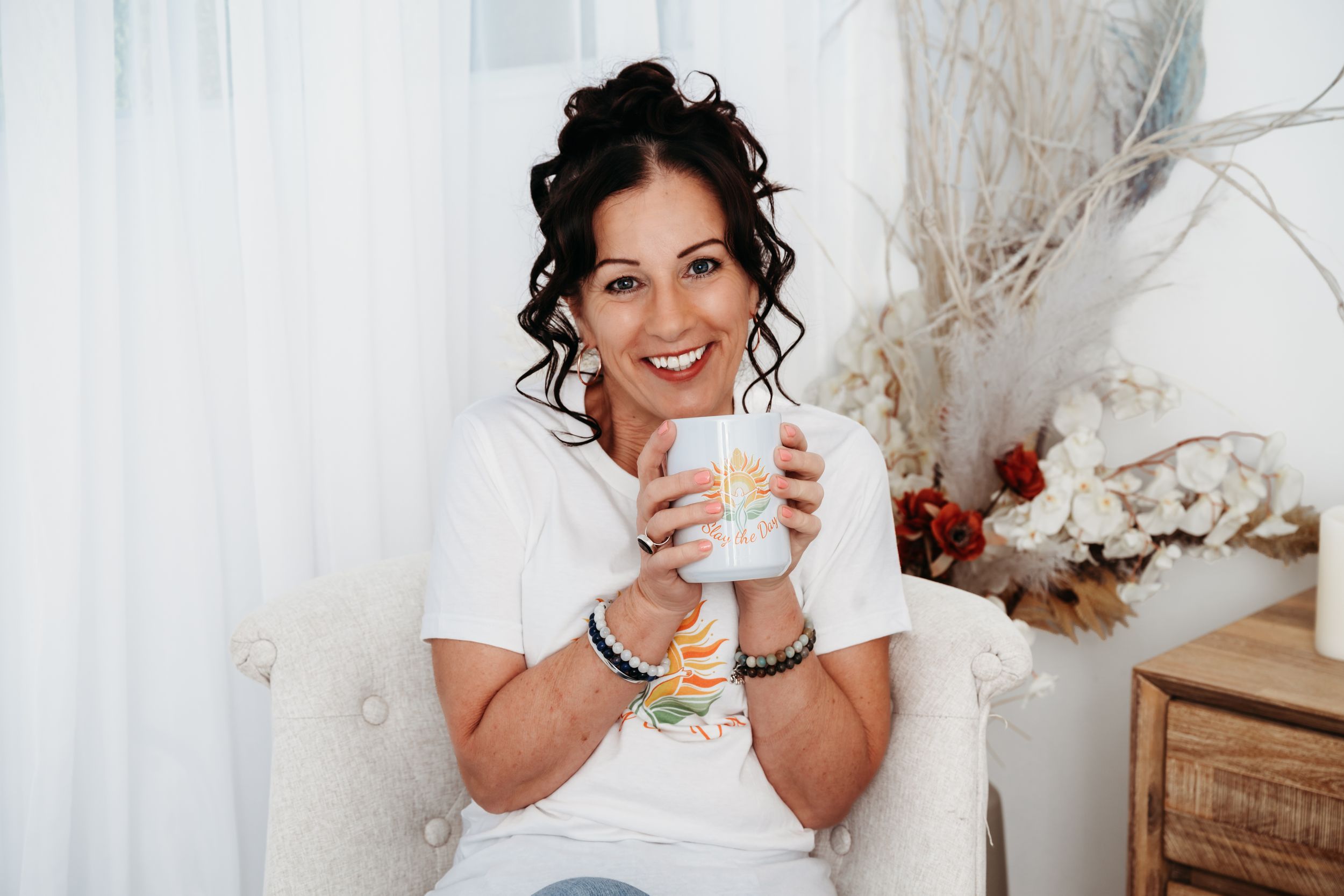 Smiling woman with curly hair holding a white mug while sitting in a cozy chair with floral decor.